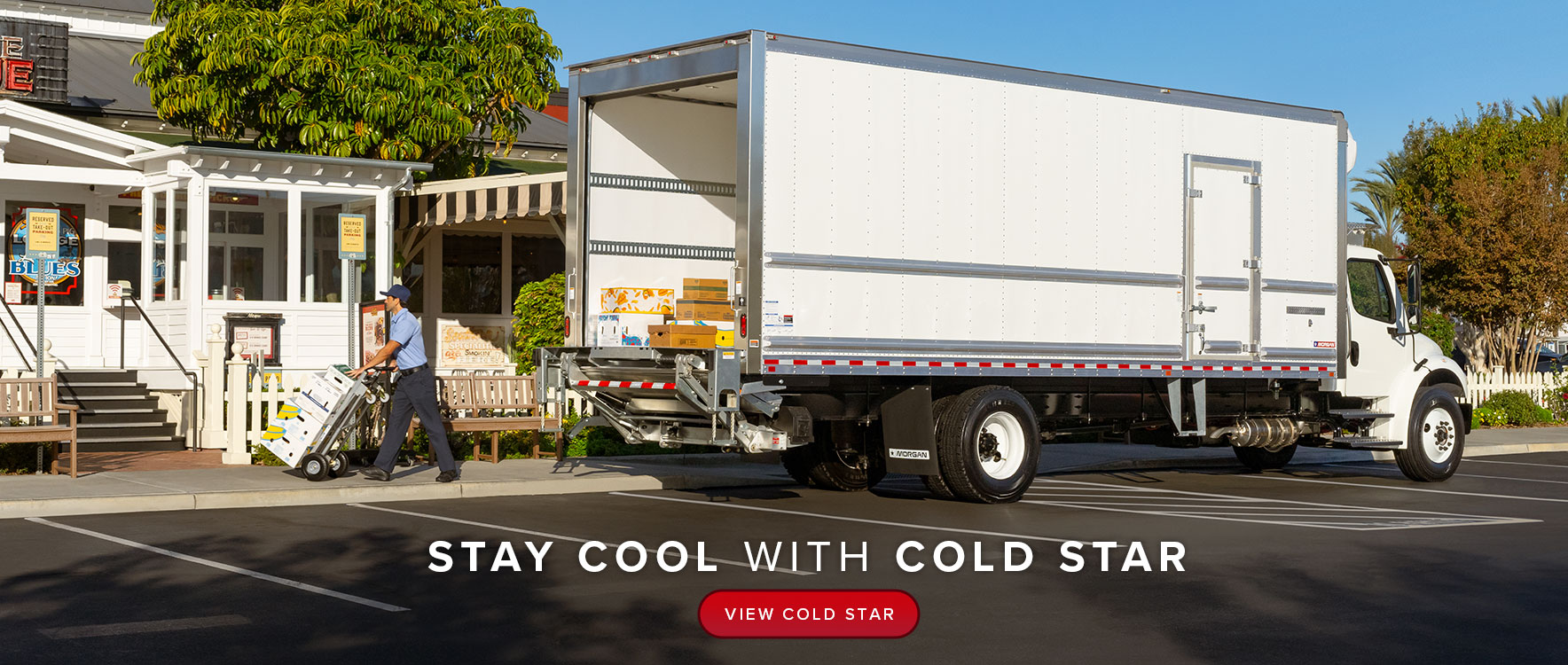 A large, white refrigerated truck is parked in front of a BBQ restaurant. A delivery man is transporting boxes of fresh produce on a dolly as he heads towards the front door.