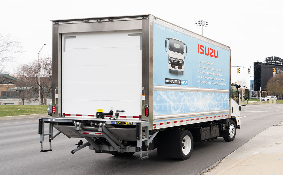 Petite benne sèche blanche avec autocollants bleus, montée sur un châssis électrique Isuzu NPR, photographiée de l'arrière sur l'autoroute.