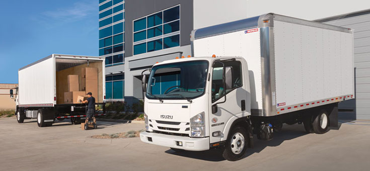 Two white dry freight trucks in a corporate setting with one truck backed into a dock while a delivery person unloads boxes from the rear of the second truck.