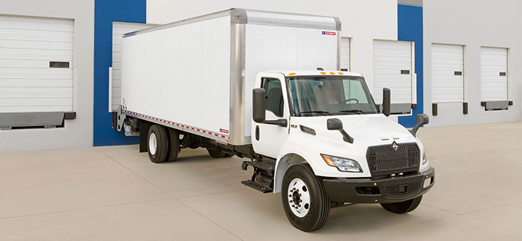 Large, white dry freight truck, backed into a loading dock at a large warehouse.